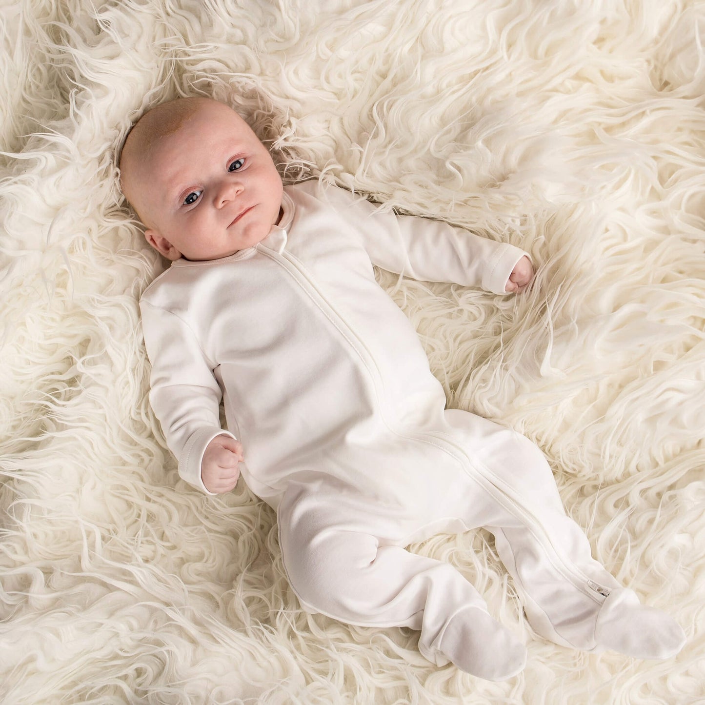 Baby less than 12 months old, laying down wearing a nueborn white double zip sleepsuit